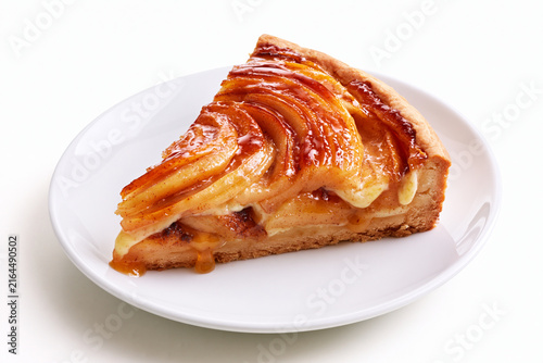 Part de tarte aux pommes sur fond blanc, sur assiette blanche. Pâte brisée, tranches de pommes caramélisées avec leur jus et migaine façon crème pâtissière