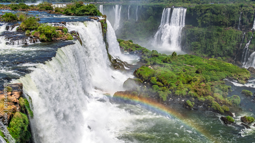 Fototapeta premium Iconic Iguazu Falls with a prominent rainbow over the churning water, captured in high resolution to highlight the mist, vegetation, and dramatic scale