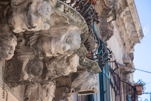 Konstfotografi Modica, Italy; 1st August 2026: Baroque architectural detail with carved grotesque face and ornate stonework on a balcony in Sicily