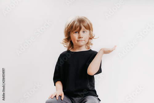 Cute little boy in black T-shirt is sitting on chair, smiling, and holding his hand near his face, showing that there's something on it.