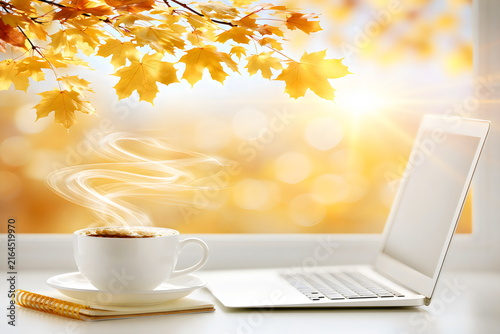 Coffee cup sits next to a laptop with golden autumn leaves in the background and sunlight shining through the window