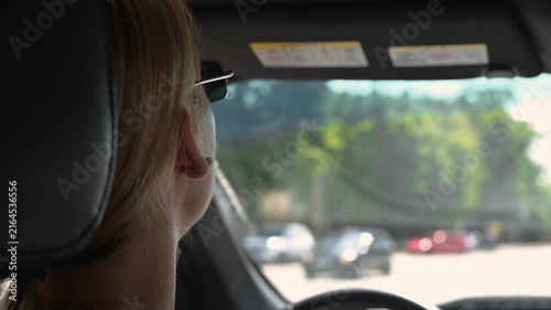 The driver sits in the front seat, wearing sunglasses, and gazes forward at the road ahead. The background shows a sunny day with trees and vehicles visible on the road