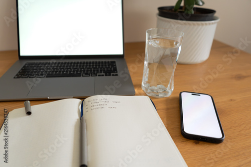 POV of minimalist office with a laptop and phone screen mockup, notebook with to-do list, glass of water and a plant on a wooden table.