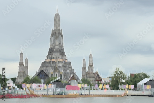 A golden Thai royal barge processional rowing past the iconic Wat Arun temple on the Chao Phraya River in Bangkok, Thailand during a traditional cultural ceremony under a bright daylight sky.