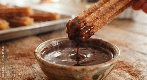 A dynamic, high-action shot of a golden churro being dipped into a small, steaming ceramic bowl of thick, glossy, dark Mexican chocolate sauce,