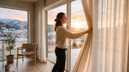 Woman opening curtains to reveal a beautiful winter sunrise over a snowy village