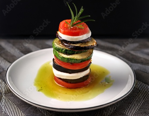 a vegetable tower on a white plate with oil and herbs the tower features layers of eggplant mozzarella zucchini and tomato all topped with melted golden cheese and a sprig of fresh rosemary