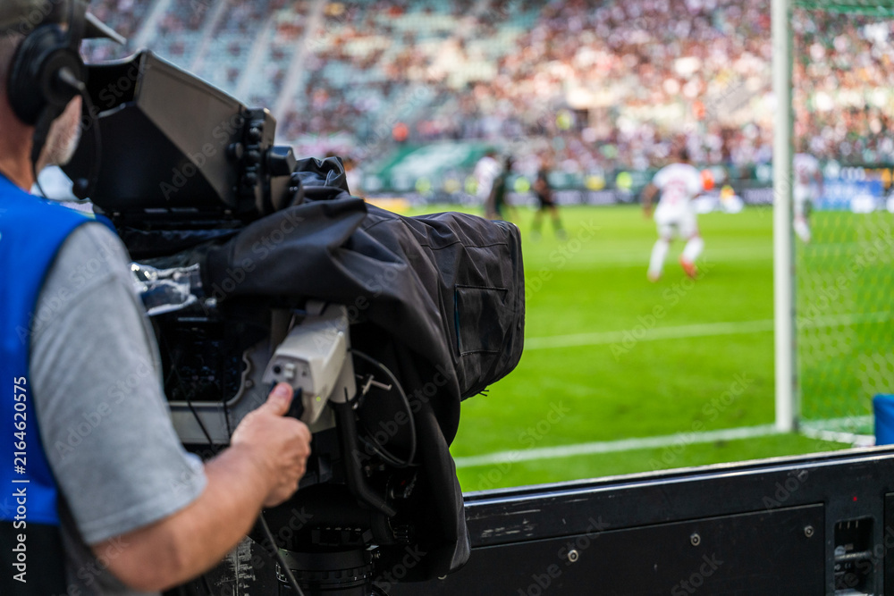 Fototapeta premium Television camera operator filming a professional soccer match from the sidelines, with a broadcast camera in the foreground and football players on the field.