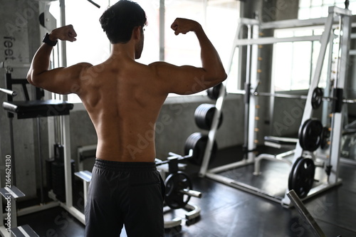 Focused muscular Asian male athlete with a sweaty bare torso standing inside a modern gym facility showing intense dedication during a heavy bodybuilding strength training workout routine.
