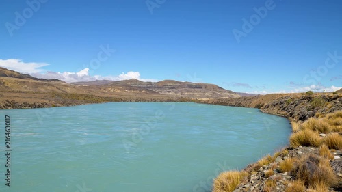 Dramatic Patagonian Steppe River: An Eye-Level Shot of the Glacial Waterway with Ripples, Flowing Towards Distant Hazy Peaks on a Sunny Day