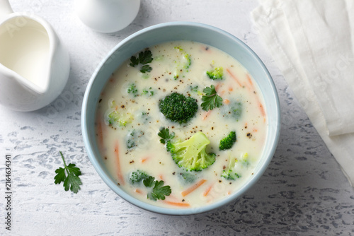 Creamy broccoli soup with potatoes, carrots and herbs in a blue bowl. Homemade vegetable soup with a rich texture on a light background