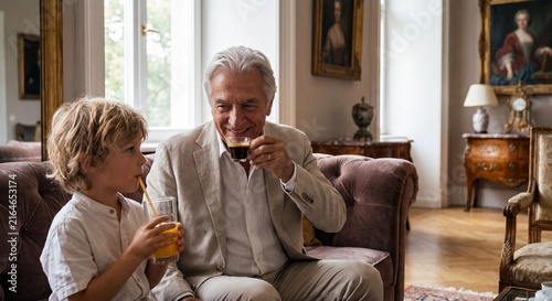 A smiling senior grandfather drinking an espresso coffee while his young grandson drinks orange juice through a straw, sitting together on a classic sofa in an elegant living room.