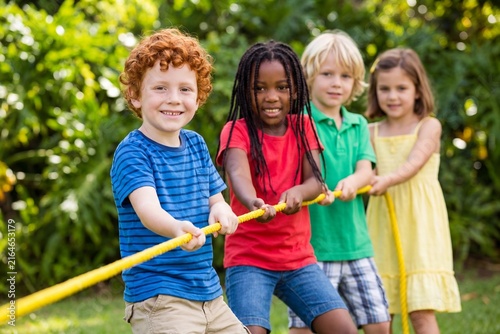 A diverse group of happy, smiling young children playing a game of tug of war with a yellow rope in a sunny, green summer park.