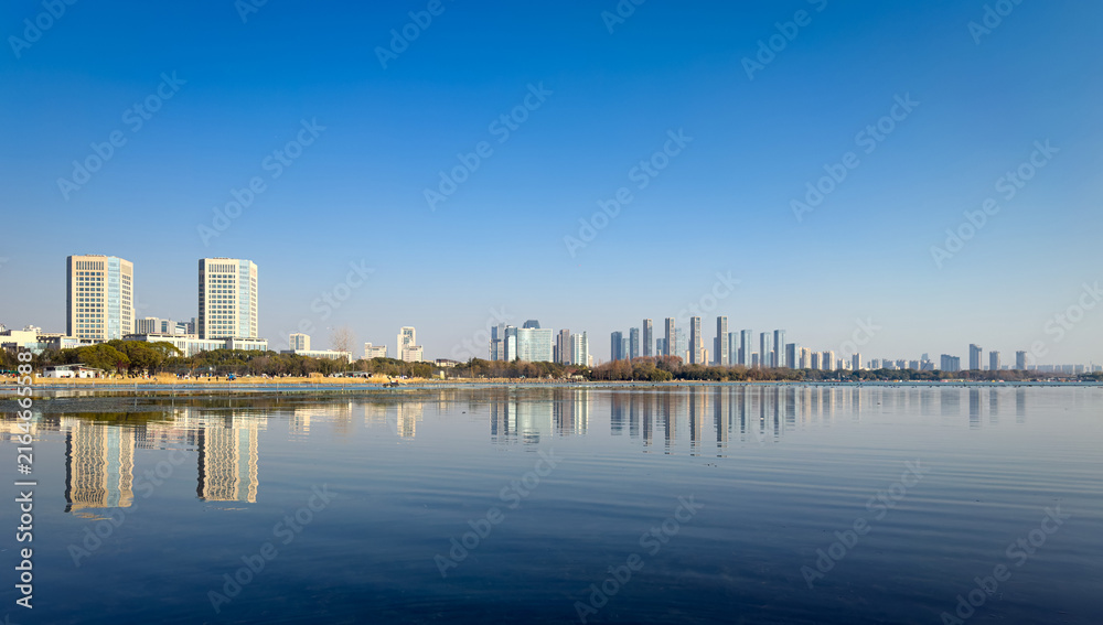 Naklejka premium Serene Waterfront Cityscape with Reflective Skyline Under Clear Blue Sky