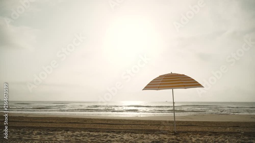Striped white orange beach umbrella stands on sandy beach in sunset sunlight against sea. Copy space background of recreational landscape without people, demonstrating calmness and serenity.