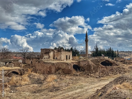 Foto Old stone village with a traditional mosque and arched ruins amid the dry winter landscape of Cappadocia, Turkey, beneath dramatic cumulus clouds and deep blue sky
