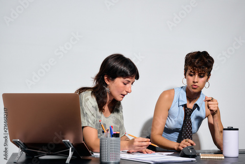 Two women collaborate intently at a desk. One writes notes while the other holds a pen thoughtfully. They share a focused, productive energy.  Direct flash. Business office,