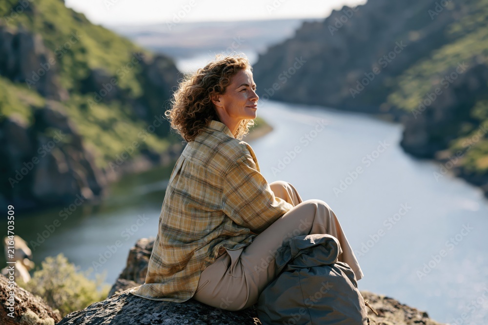 Fototapeta premium Woman sitting on cliff overlooking a river canyon for hiking travel background and outdoor adventure wallpaper with golden sunlight