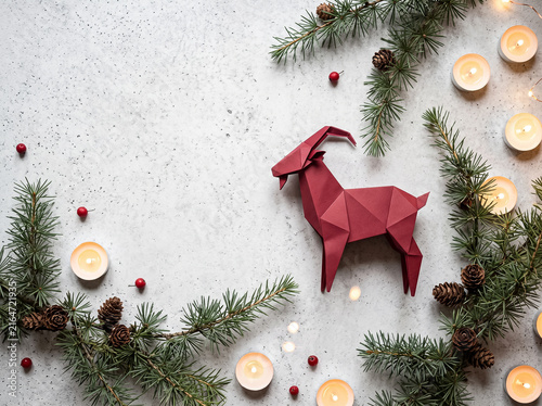 red paper goat figurine in center of composition made of fir branches cones berries and candles on light background holiday flatlay