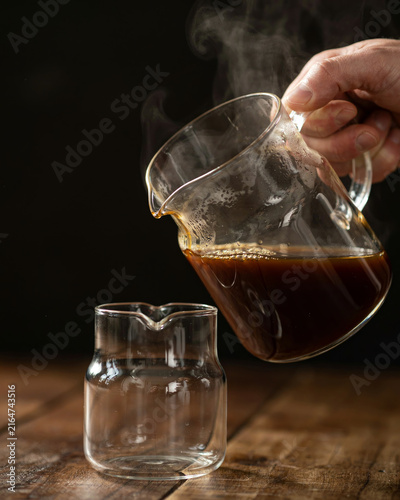 Pouring hot coffee from a glass pot into a cup on a wooden table with beverage