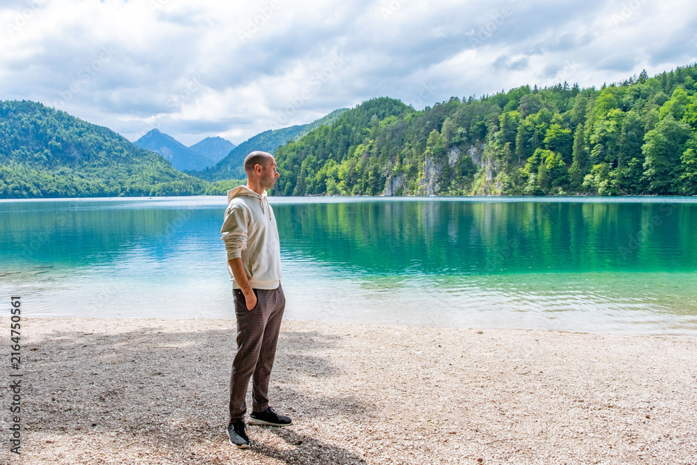 Fototapeta premium Bald young man wearing glasses, white hoodie, plaid trousers, Travel tourism stylish clothing choices, pebble beach, turquoise lake, mountains, forest trendy attire, summer vacation, fashion portrait