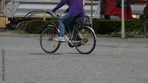 Fahrradfahrerin mit lila Pullover und Korb auf einem Citybike beim Überqueren eines großen Platzes im urbanen Stadtverkehr mit Baustellenfahrzeugen im Hintergrund – Leipzig, Sachsen, Deutschland