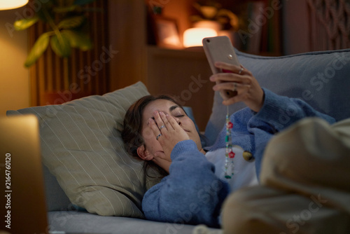 Foto Tired young adult Caucasian woman lying on sofa, holding smartphone and covering