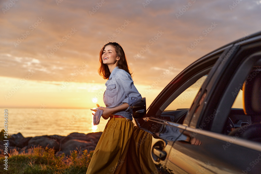 Fototapeta premium Woman lifestyle portrait by a car at sunset, smiling as she steps away, coastal landscape, warm golden hour light, casual fashion, relaxed mood, vibrant sky