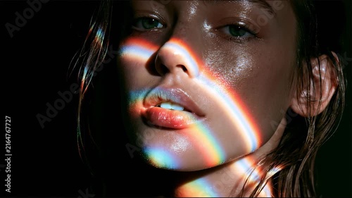 Close-up beauty portrait of a young woman as moving rainbow light reflections illuminate her face, creating a vibrant artistic fashion scene with colorful cinematic lighting