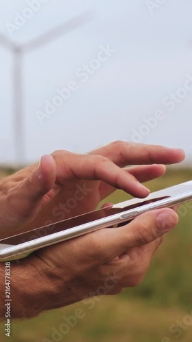 Close-up of man's hands using tablet outdoors with wind turbines in background. Digital technology and renewable energy highlight clean power, innovation, sustainability and energy management.