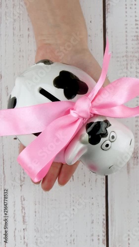 Cute ceramic cow piggy bank with pink ribbon held by hand. Savings concept illustrates financial education, money management and saving habits for children in playful home setting.