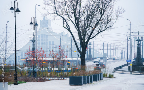 Cuadro en lienzo Winter urban transition appears where bare trees, tram lines and a modern glass building recede into mist near Riga Old Town, supporting city mobility and seasonal weather stories