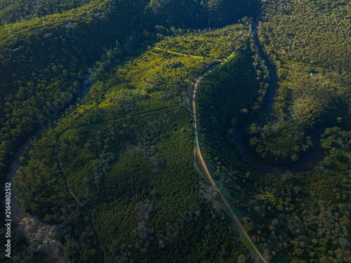 Aerial view of a winding river flowing through a dense forest with a dirt path cutting through the lush green landscape in Wonga Park, Victoria, Australia.