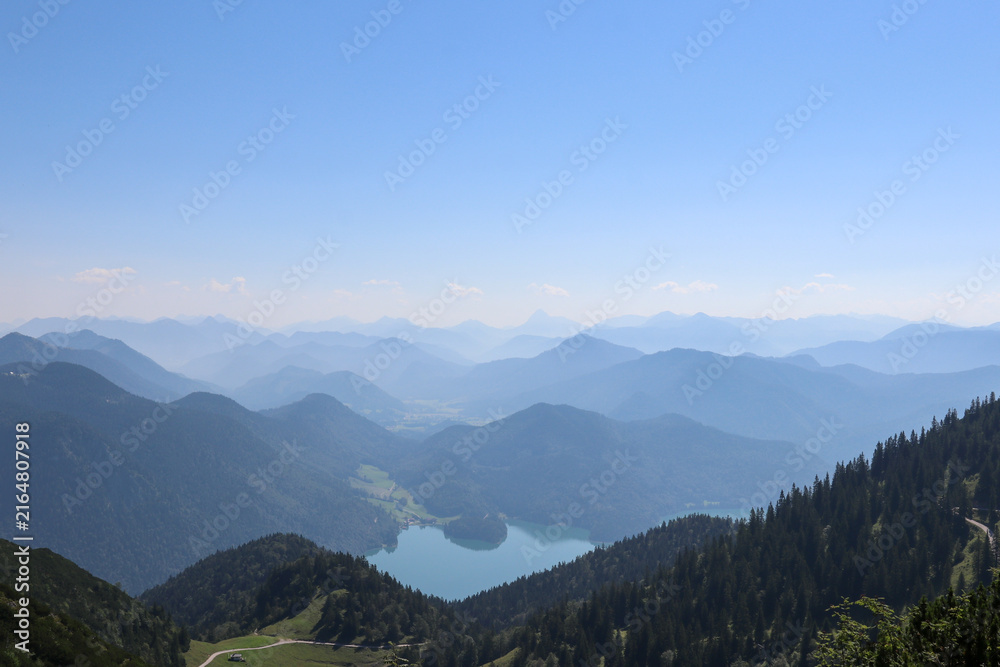 Fototapeta premium Atemberaubender Weitblick von einem Berggipfel auf einen tiefblauen, eingebetteten Bergsee, umgeben von dichten Nadelwäldern und gestaffelten Alpenketten im Dunst.
