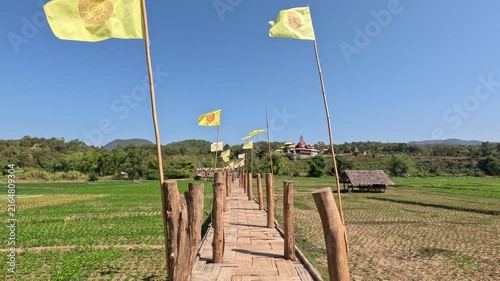 Wide shot of an old bamboo bridge at noon in northern Thailand, featuring Buddhist temple flags and rice paddies along the path leading to a distant temple under a clear blue sky.