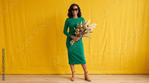 Stylish woman in green dress holding dried flowers against yellow background