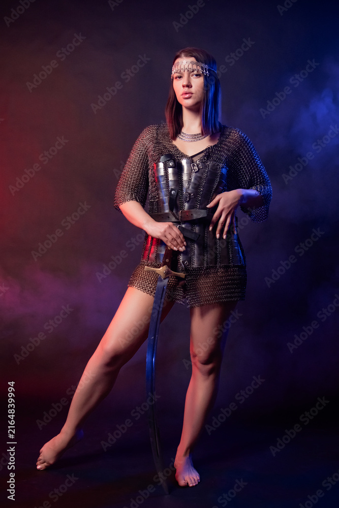 Fototapeta premium A portrait of a young woman in oriental chainmail armor, holding a saber and posing standing against a dark background with red and blue backlighting. Concept art of an oriental warrior.