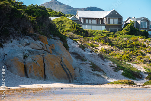 Konstfotografi Modern residential houses sit on a vegetated sandy hillside above granite boulders and shallow water in Cape Town, South Africa