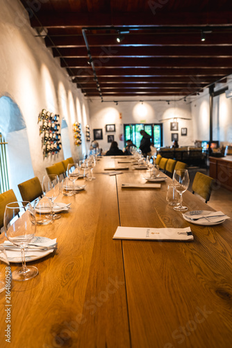 Bild auf Leinwand Wine glasses and place settings rest on a long wooden dining table inside a Cape Town winery tasting room with timber ceilings and seated visitors