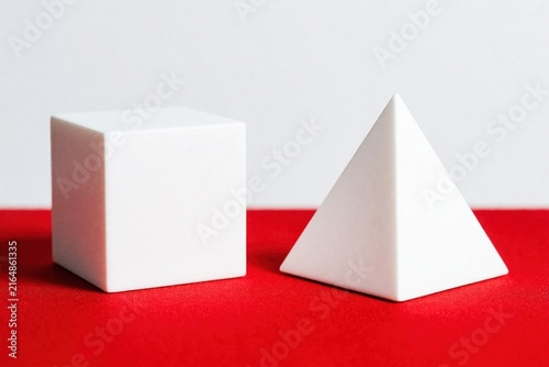 Obraz na płótnie White cube and triangular pyramid stand on a red surface against a grey backdrop. Simple geometric forms display clean lines and solid shapes, offering abstract composition.