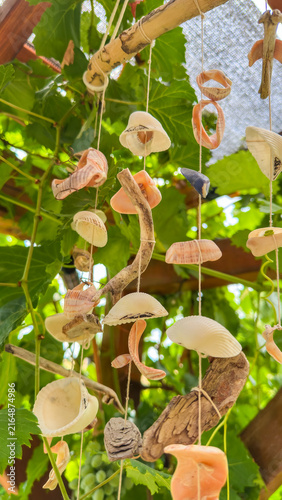Konstfotografi Handmade seashell wind chime hangs from a wooden pergola amidst green leaves