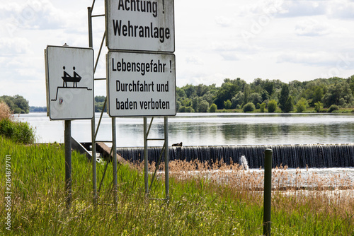Warning signs near a river indicating danger and prohibitions against swimming and navigation, with a weir and lush greenery in the background