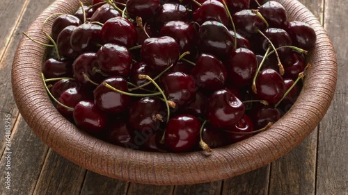 Sweet cherries with stems on a handmade ceramic plate. Fresh organic fruits on a vintage ribbed dish with a weathered, dark rustic wood background, table spin, backlit natural light.