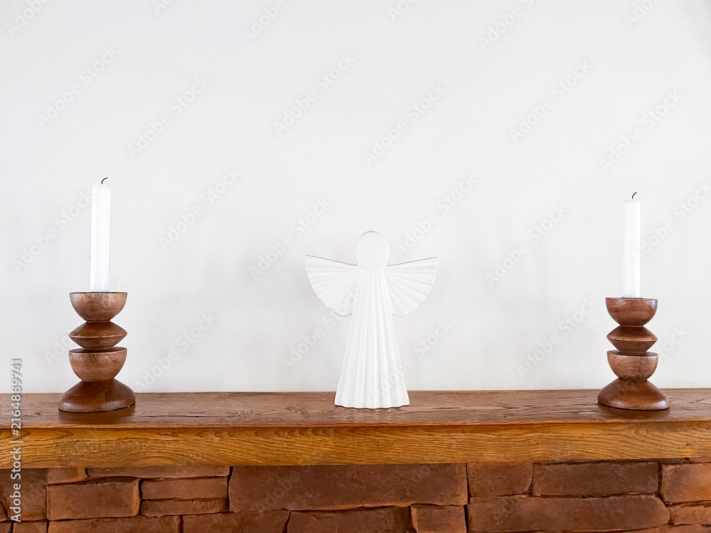 Fototapeta premium White Angel Figurine and Candles on Wooden Shelf Above Stone Fireplace
