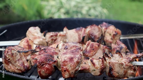 Marinated pieces of meat grilling on metal skewers over a hot charcoal grill in a summer garden with fresh green grass in the background. Close-up barbecue scene