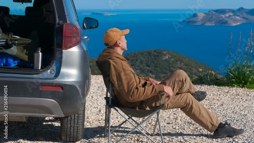 Traveler rests near a car. Relaxed man sitting in a camping chair next to his car on a cliffside, enjoying the stunning panoramic view of the blue sea and islands during a peaceful road trip break