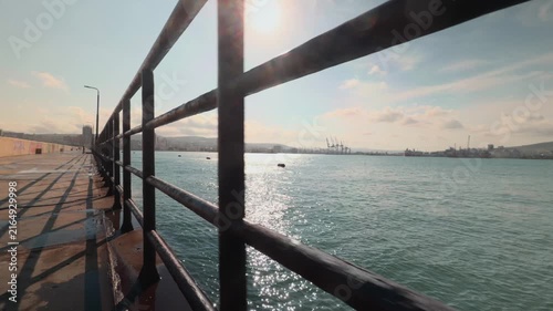 Rusty metal railings on concrete pier at maritime port city harbor with sun flare and water reflection during sunny daytime wide shot tracking forward to reveal distant cargo ships and skyline
