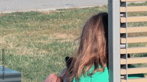 Unrecognizable female digital nomad wearing green shirt sitting on wooden bench outdoors using mobile device. Wind blows long hair close up rear view mindful break park green grass sunny day