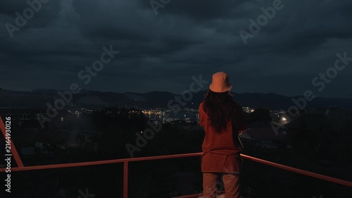 Unrecognizable anonymous long haired man in bucket hat standing on terrace with crossed arms then putting hands behind head looking at night coastal city port lights and cloudy sky rear view static