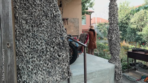 Unrecognizable long haired man in bucket hat entering frame and walking away along concrete porch disappearing around corner of rustic rural house with camouflage netting in summer garden rear view